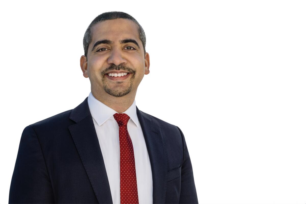 A photo of a smiling man with a goatee and mustache wearing a dark blue suit, white collared shirt and red tie