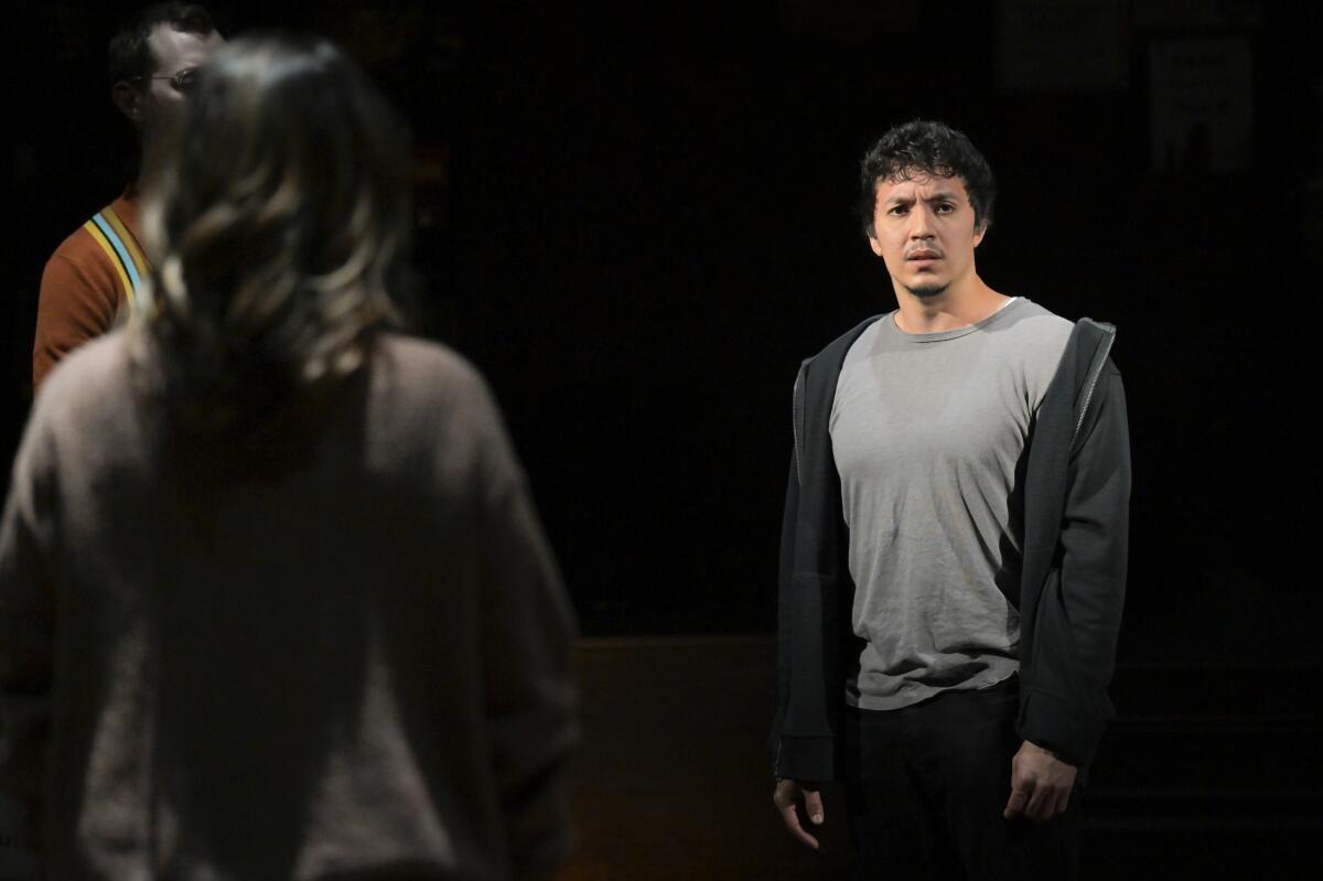 A man in a gray T-shirt and hoodie stands on a darkened stage with two other people.