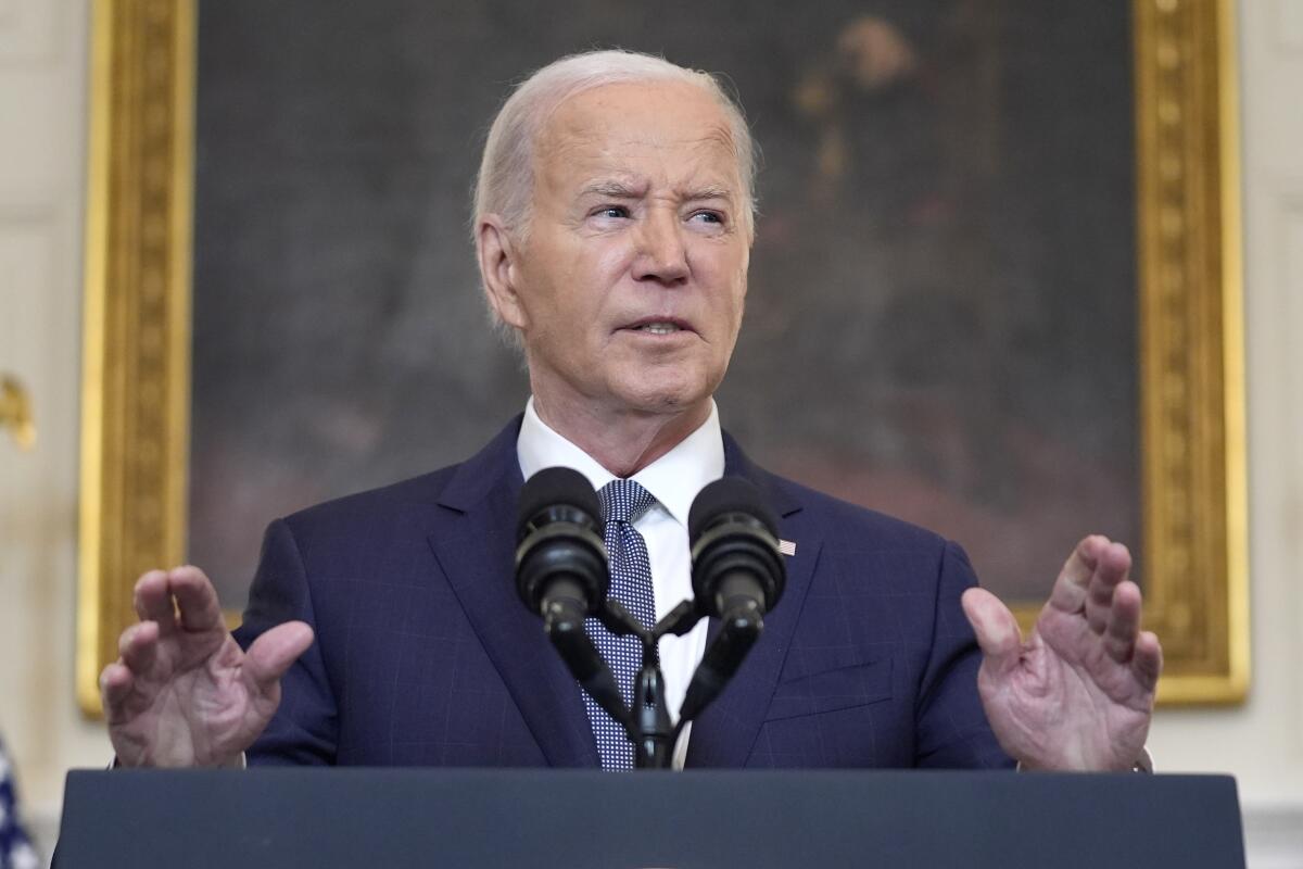 Joe Biden, wearing a dark suit and blue tie, gestures with his hands while standing at a lectern 