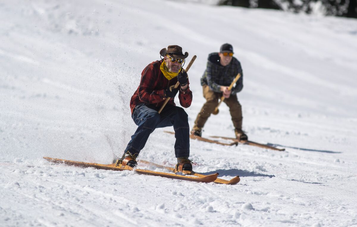 Two men wearing plaid shirts on wooden skis glide down a snowy hill.