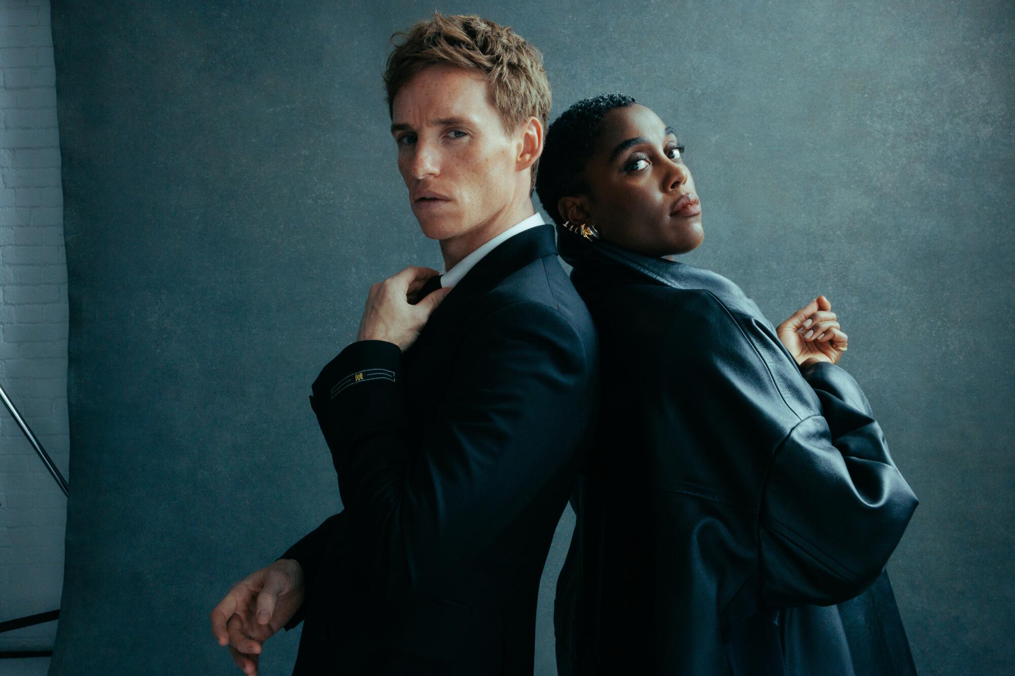 Eddie Redmayne and Lashana Lynch standing back to back in front of a gray backdrop.