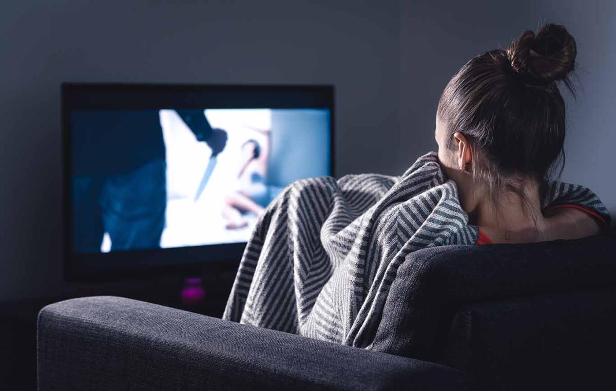 Scared woman watching horror movie on tv hiding under blanket on couch at night.