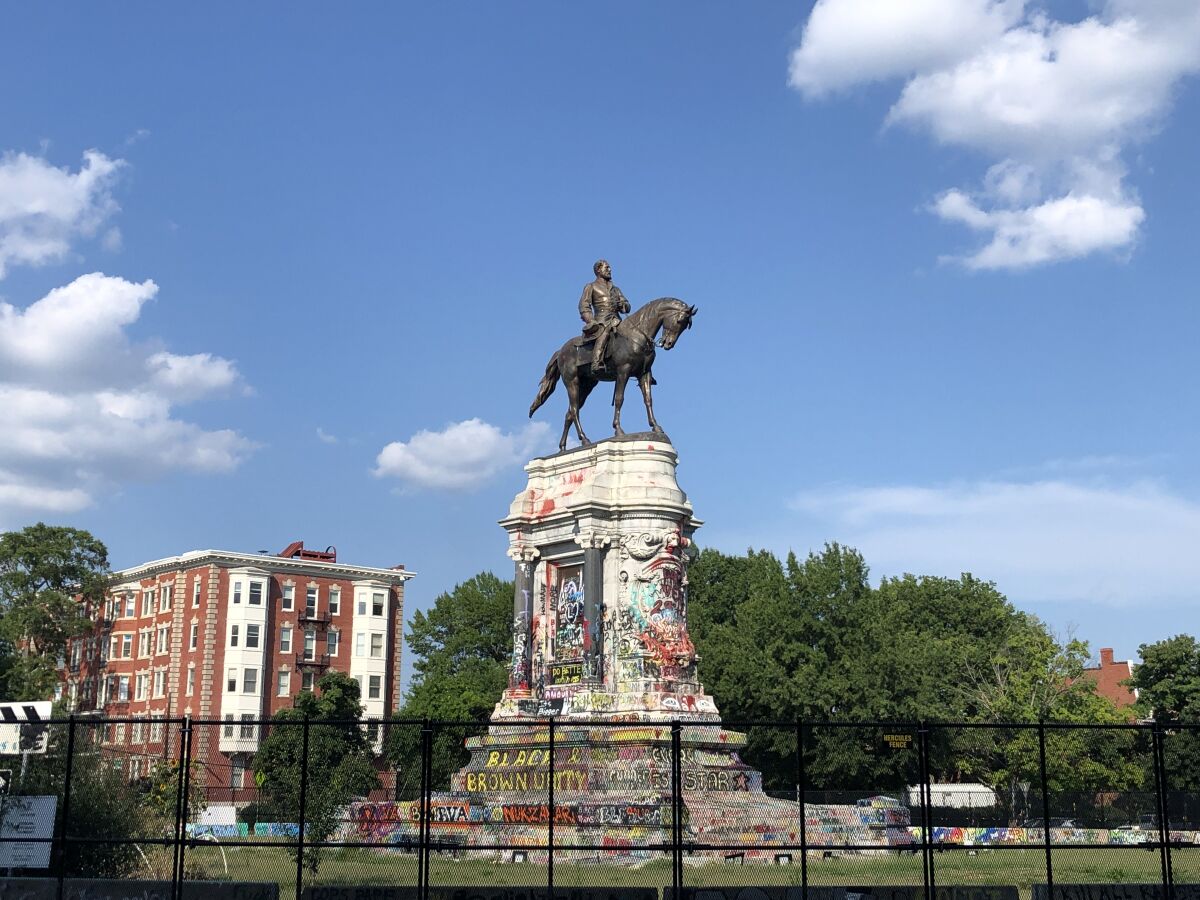 The Robert E. Lee Monument is shown surrounded by a fence and covered in colorful graffiti