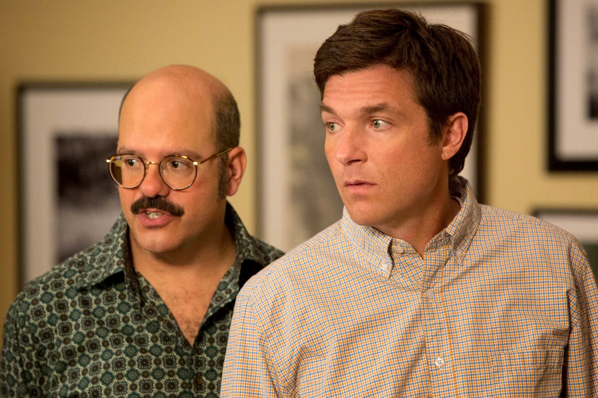 A man in a green patterned shirt and glasses stands beside a man in a button-down plaid shirt.