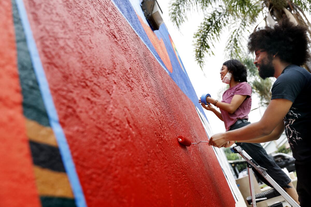 Two people on ladders paint a mural on the outside of a building