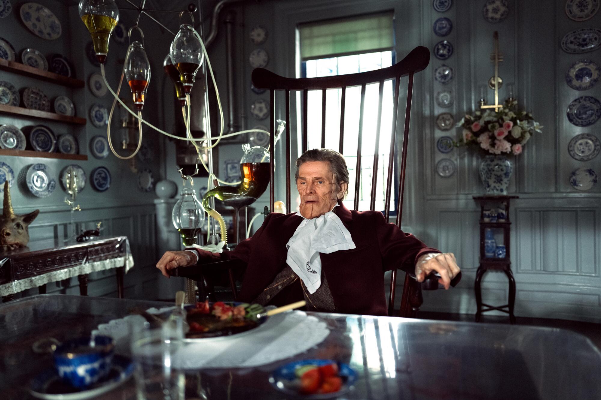 A man with severe facial scarring sits in a large chair in a scene from "Poor Things."