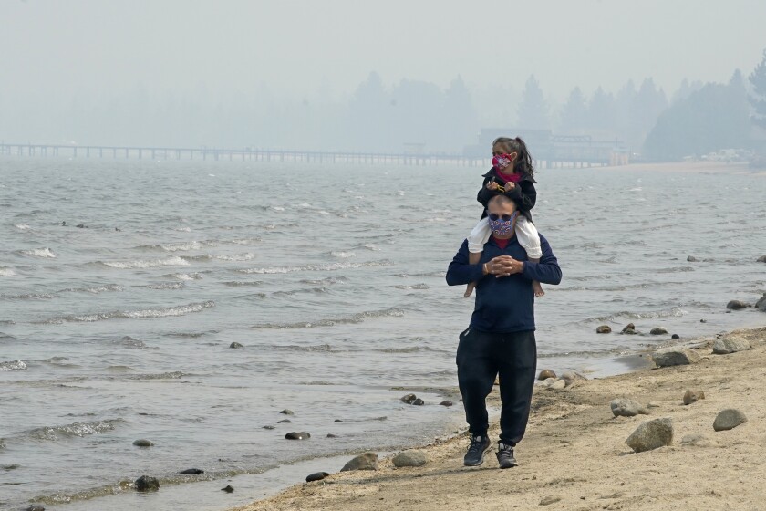 Presley Mila Perez gets a ride on the shoulders of George Estrada as they walk along the shore of Lake Tahoe in South Lake Tahoe, Calif., Tuesday, Aug. 24, 2021. The pair both wore face mask to protect them from the smoke from the Caldor Fire that is blanketing the area cloaking the normally blue sky in a dull gray aura. (AP Photo/Rich Pedroncelli)