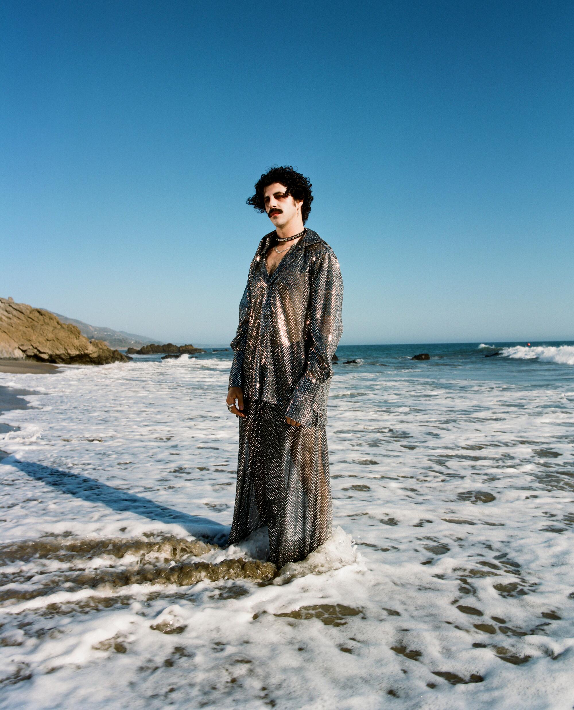 A man in a long glittery outfit stands in the ocean.