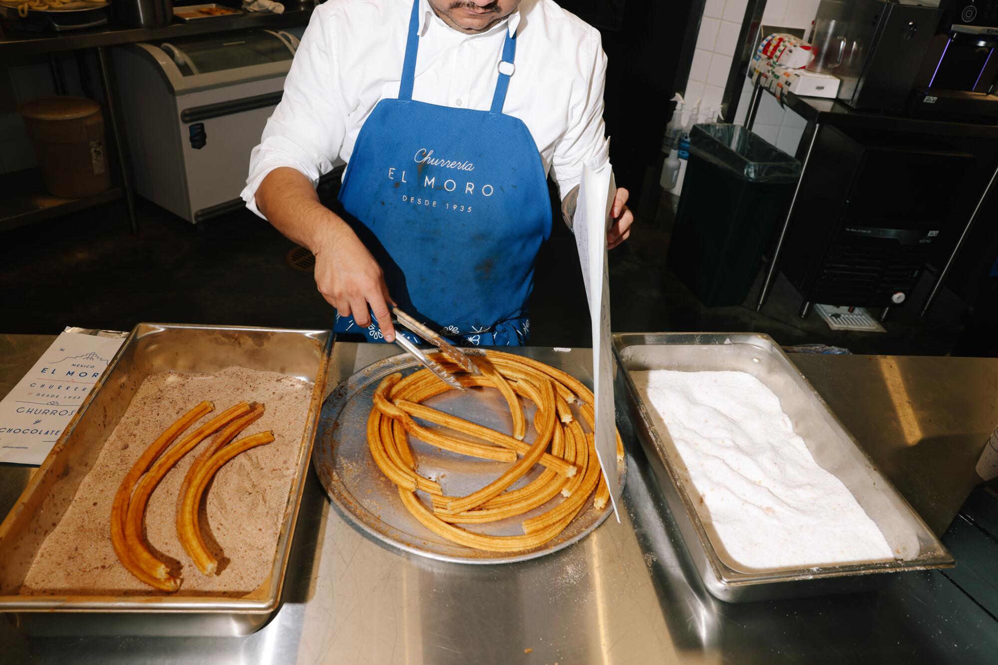 A high-angle view of someone in an apron using tongs to jostle churros on a round platter. Sugar sits in a tub on the side.