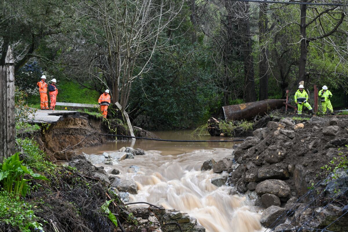 Several utility workers watch from the banks as a brown, overflowing creek cuts a road in half.
