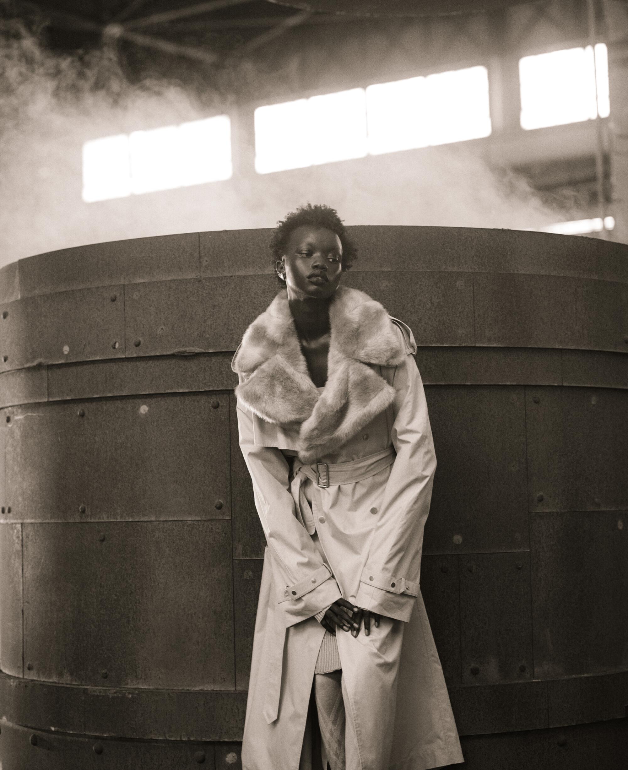 Sepia-toned photo of a model in a trench coat in front of a vat that appears to be steaming