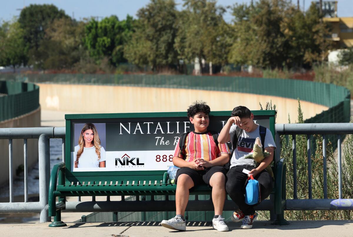 Two teen boys sit at a Metro bus stop in the full sun.