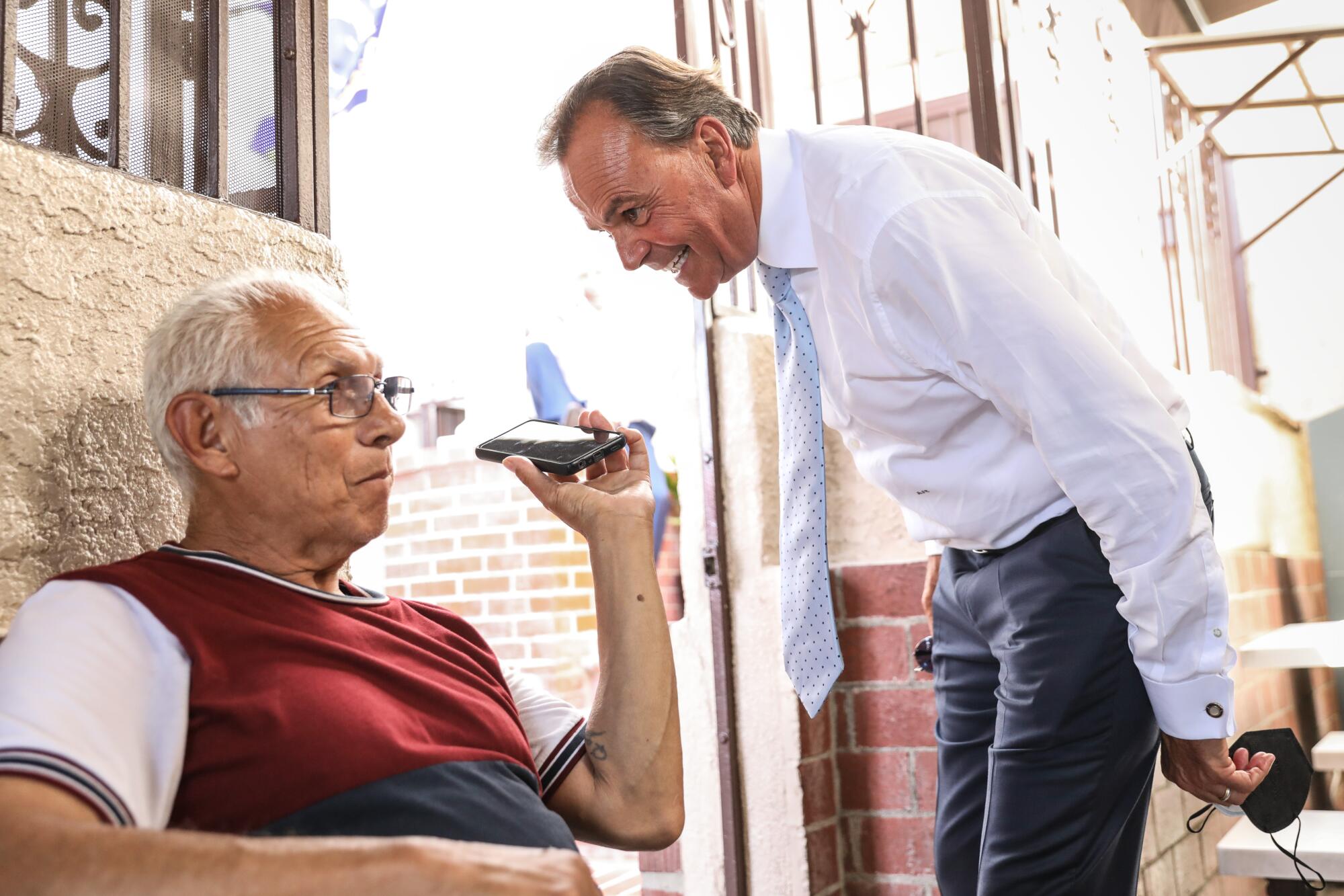 Rick Caruso standing and leaning down to talk on a cellphone held up by a seated man 