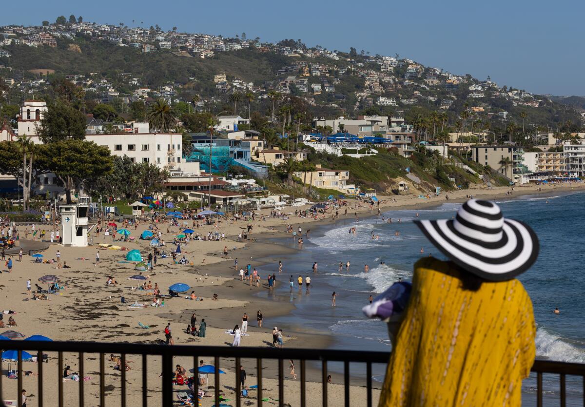 A person in a black-and-white striped hat and orange top stands at a railing looking at people on the beach below