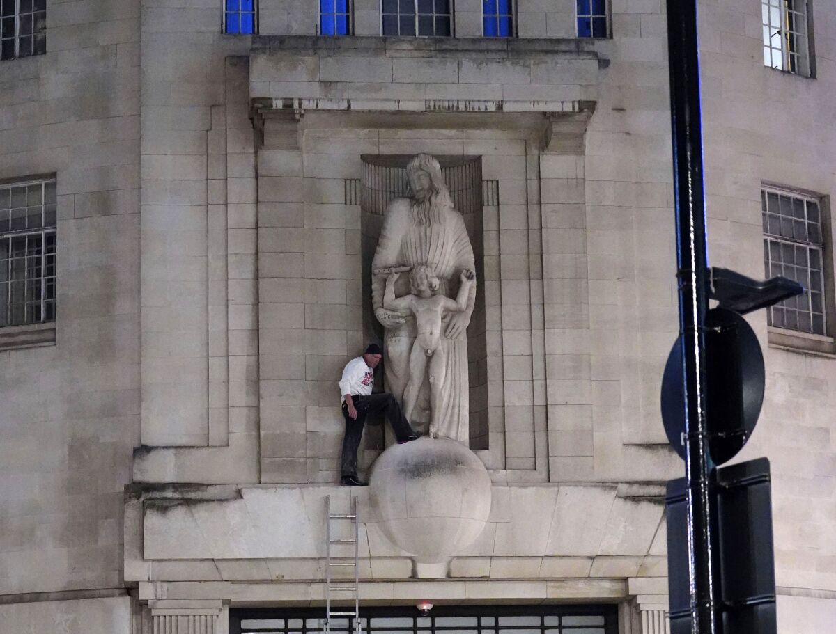 A man, after climbing a ladder, stands next to the statue of Prospero and Ariel from Shakespeare's play The Tempest by the sculptor Eric Gill outside the BBC's Broadcasting House in central London, Wednesday Jan. 12, 2022. British police have cordoned off an area outside the BBC in central London after a man was spotted scaling the building and using a hammer to attack a statue by controversial sculptor Eric Gill. (Ian West/PA via AP)