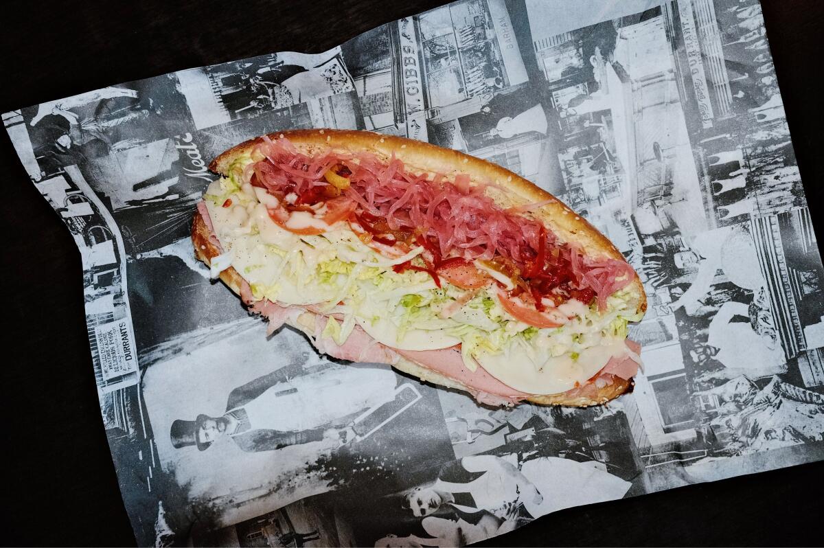 A long, full-size sandwich, fully loaded, with sliced ​​meats, cheese, and pickled onions on black and white paper at Gwen.