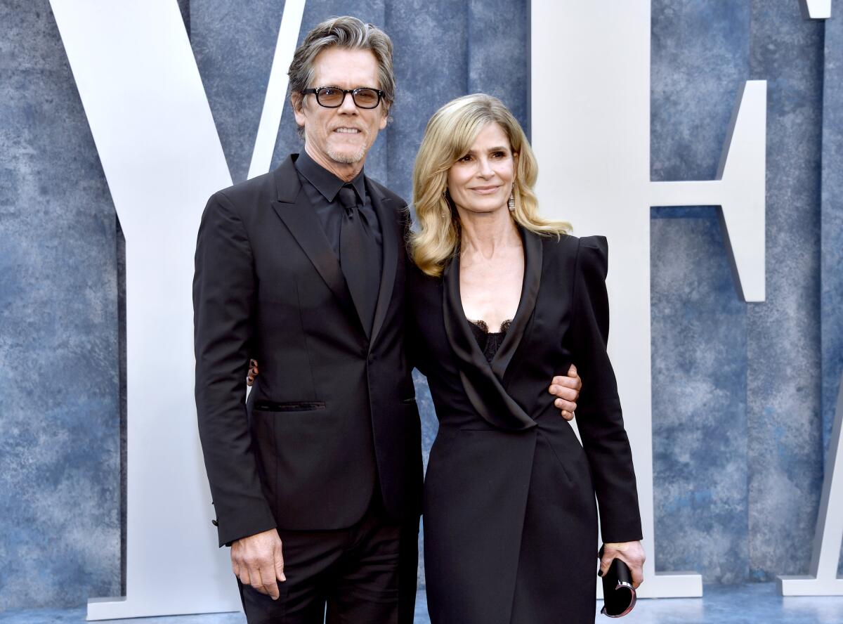 Kevin Bacon wears an all-black suit and Kyra Sedgwick wears a black dress as they stand arms around each other