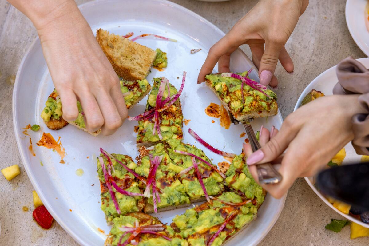 Hands pick up pieces of avocado toast from an oval plate.