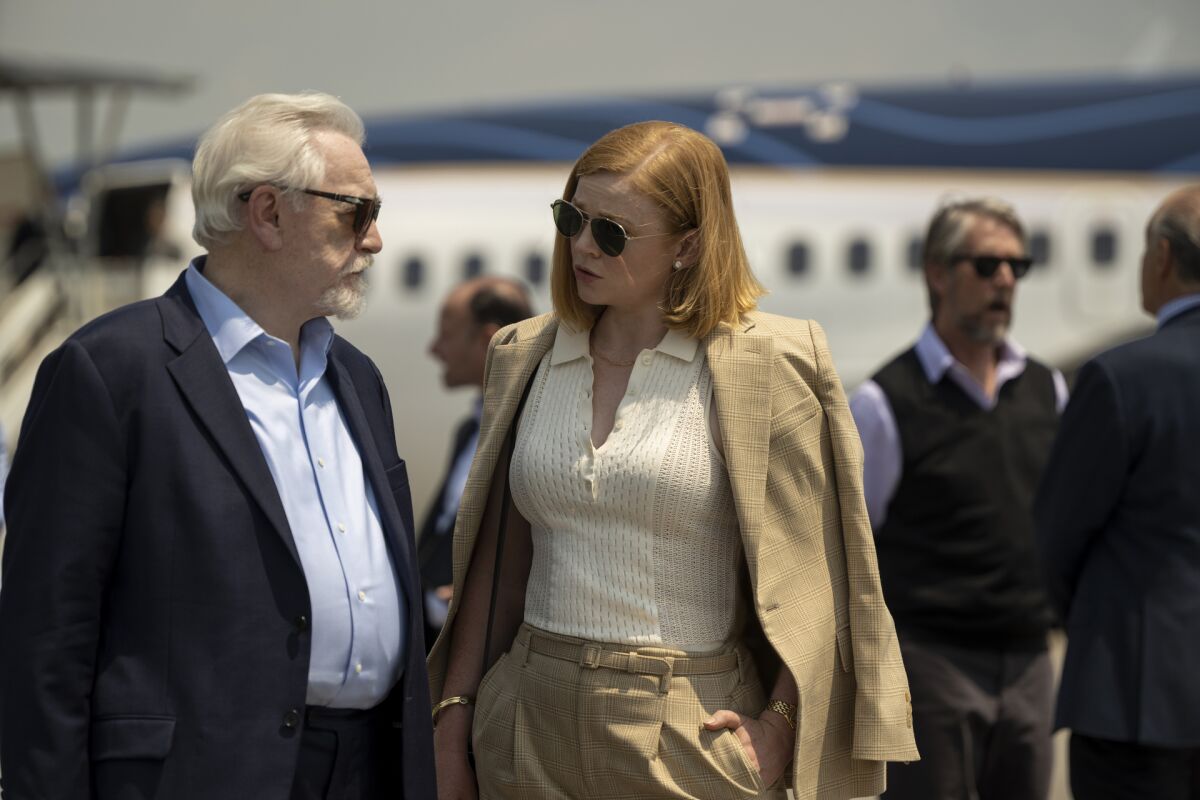 A man and a woman wearing suits and sunglasses talk