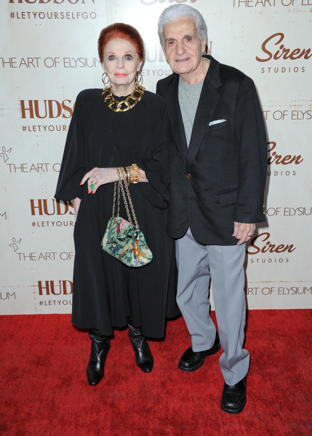 A woman with red hair in a black dress stands next to a man in a black jacket and gray pants on a red carpet.