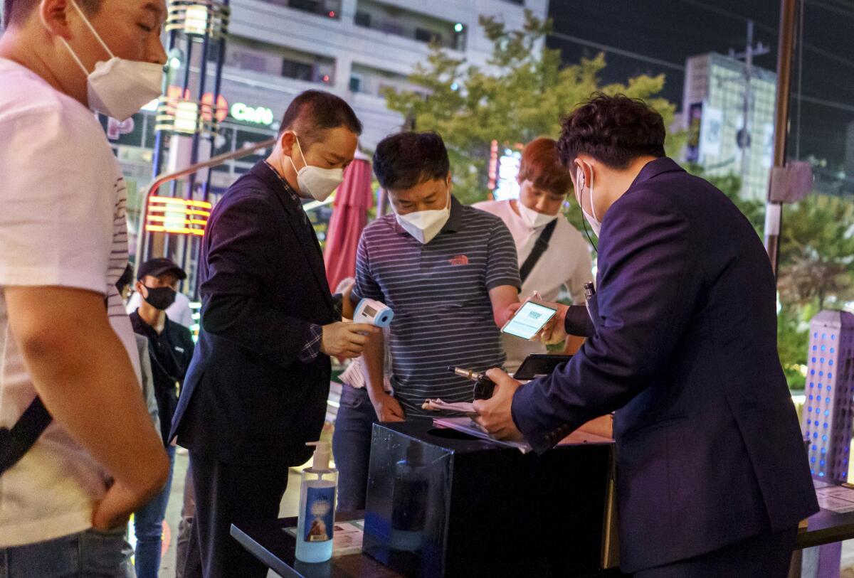 A man in a line shows his phone to two other men, one of whom holds a forehead thermometer.