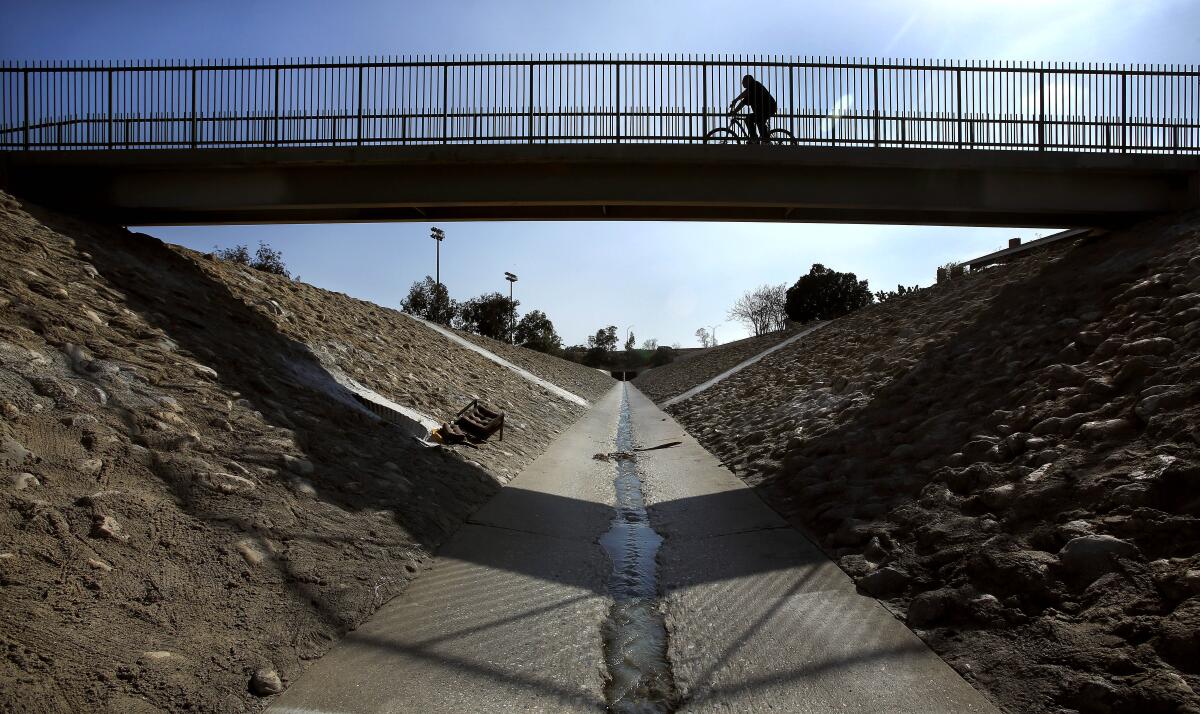 A trickle of water flows along a concrete channel beneath a bridge on which a bicyclist rides.