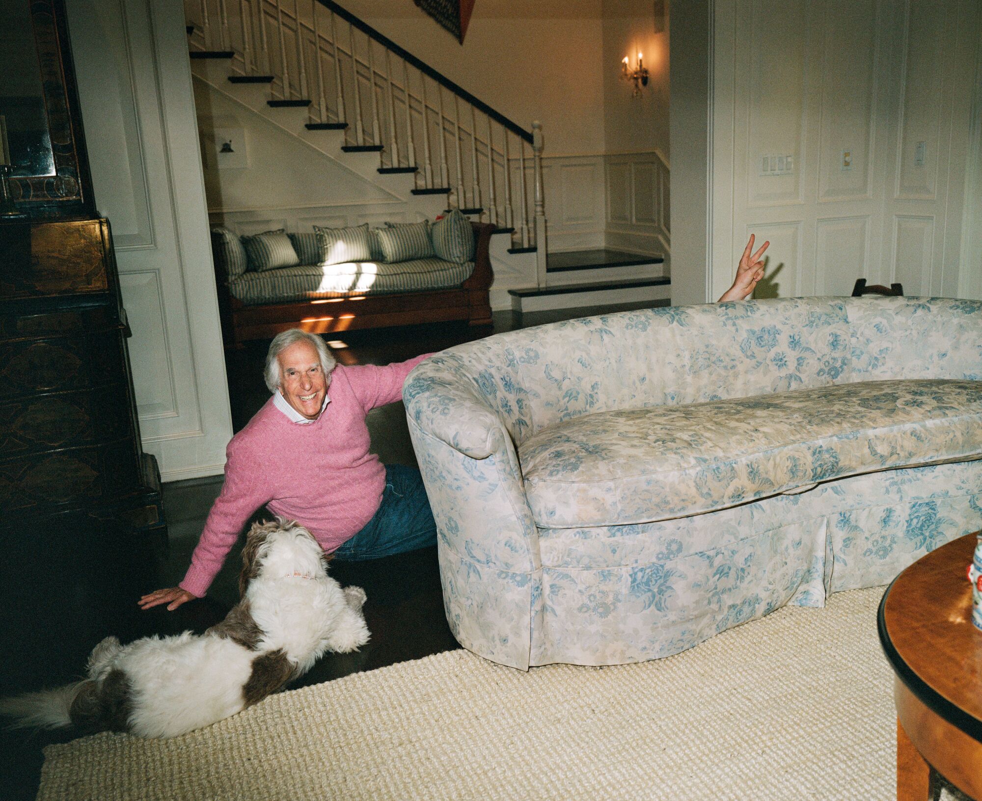 A man with a pink sweater sits on the ground with a dog as a hand is seen from a behind a floral couch