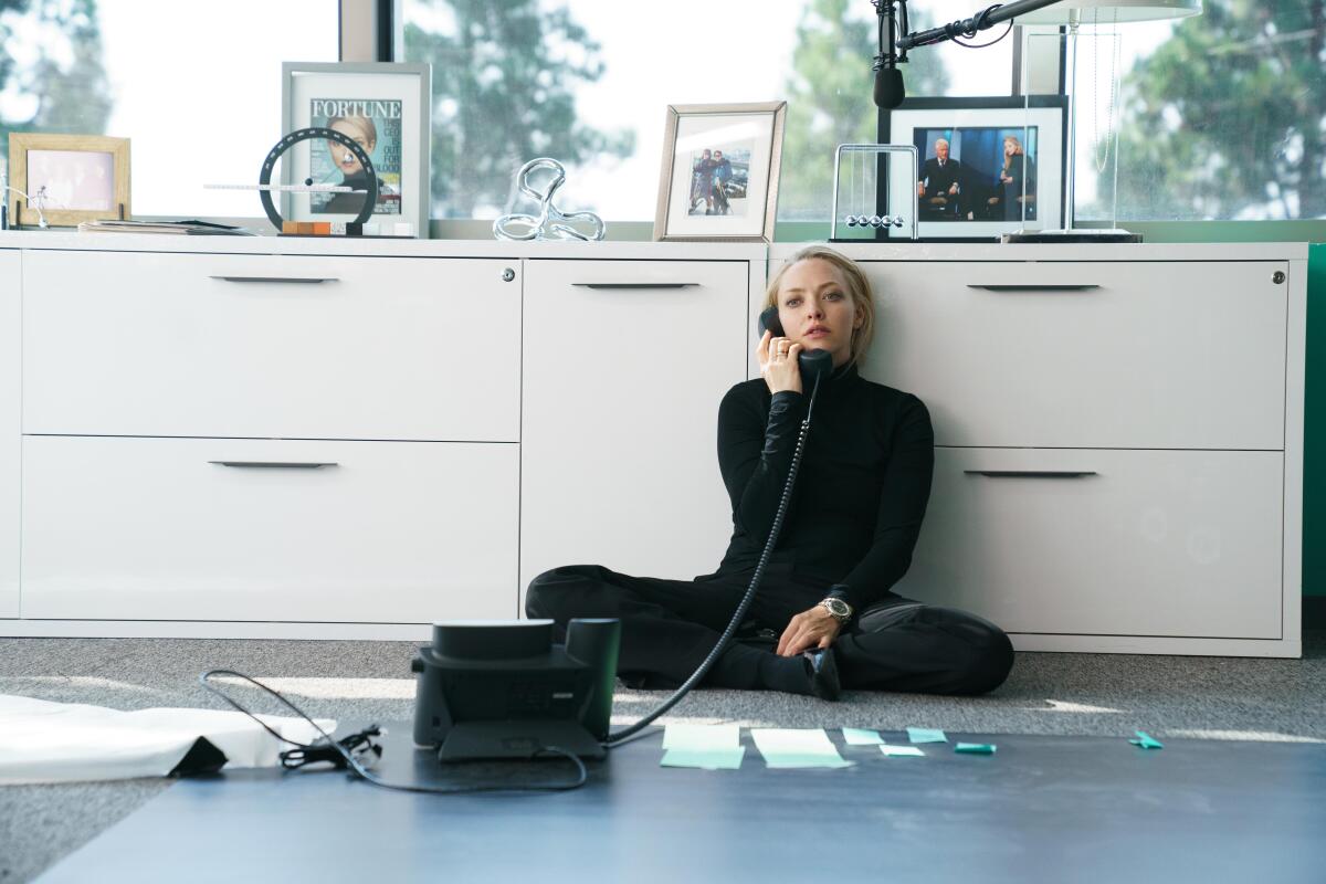 A woman dressed in black sits on the floor, leaning against a white dresser, talking on the phone.