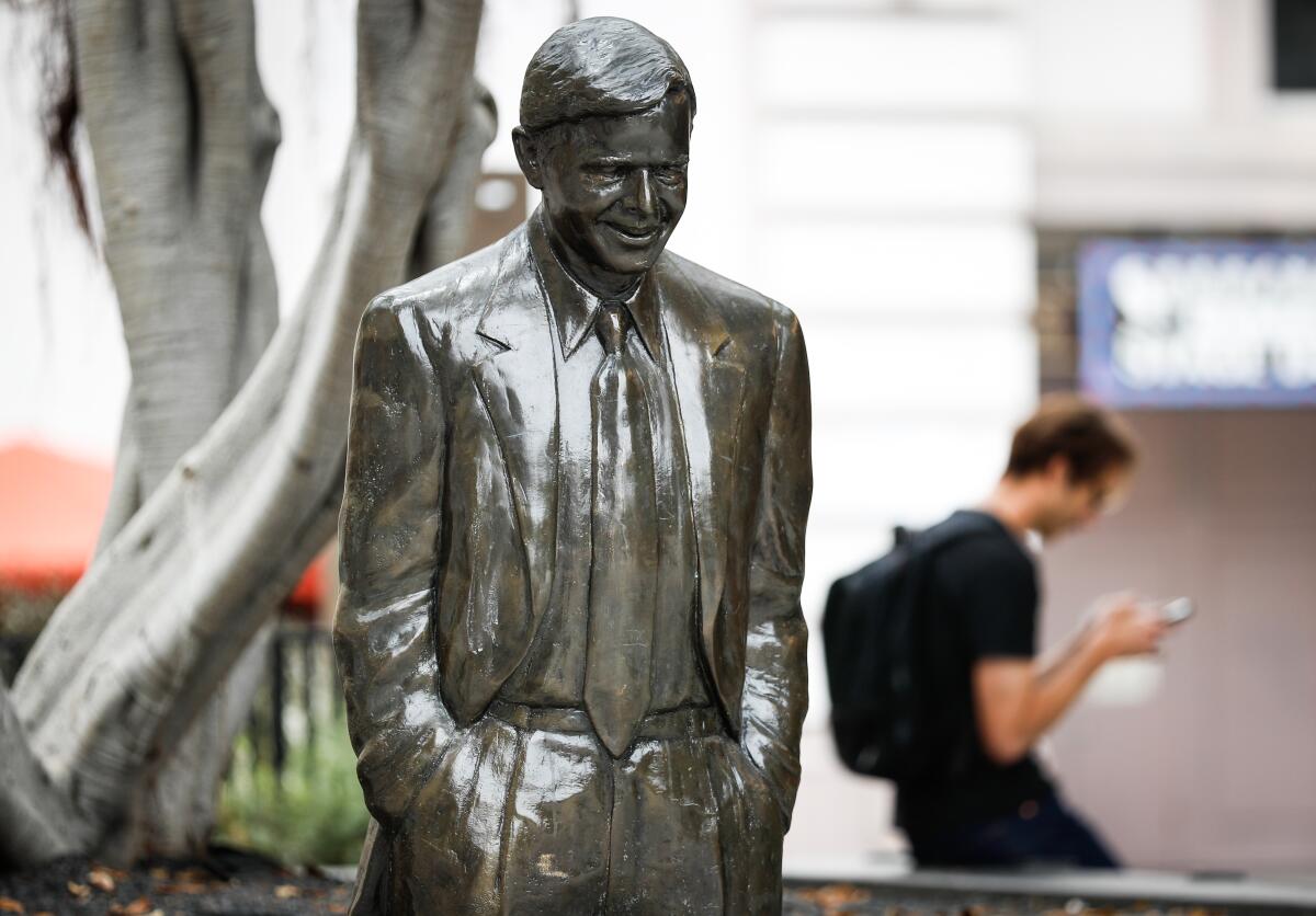 A bronze statue of a man in a suit is pictured on a sidewalk, as a man in a backpack looks at his cell phone.