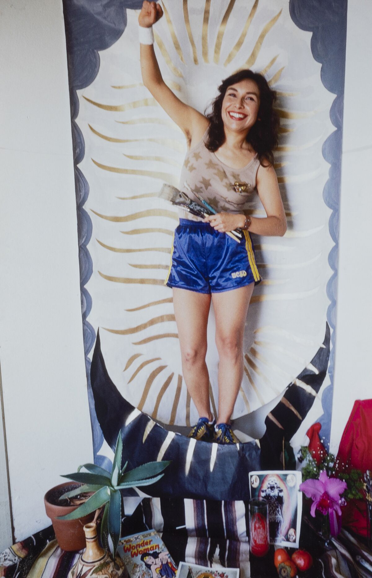 A woman  in running shorts and holding paintbrushes stands before a backdrop typical of the Virgin of Guadalupe.
