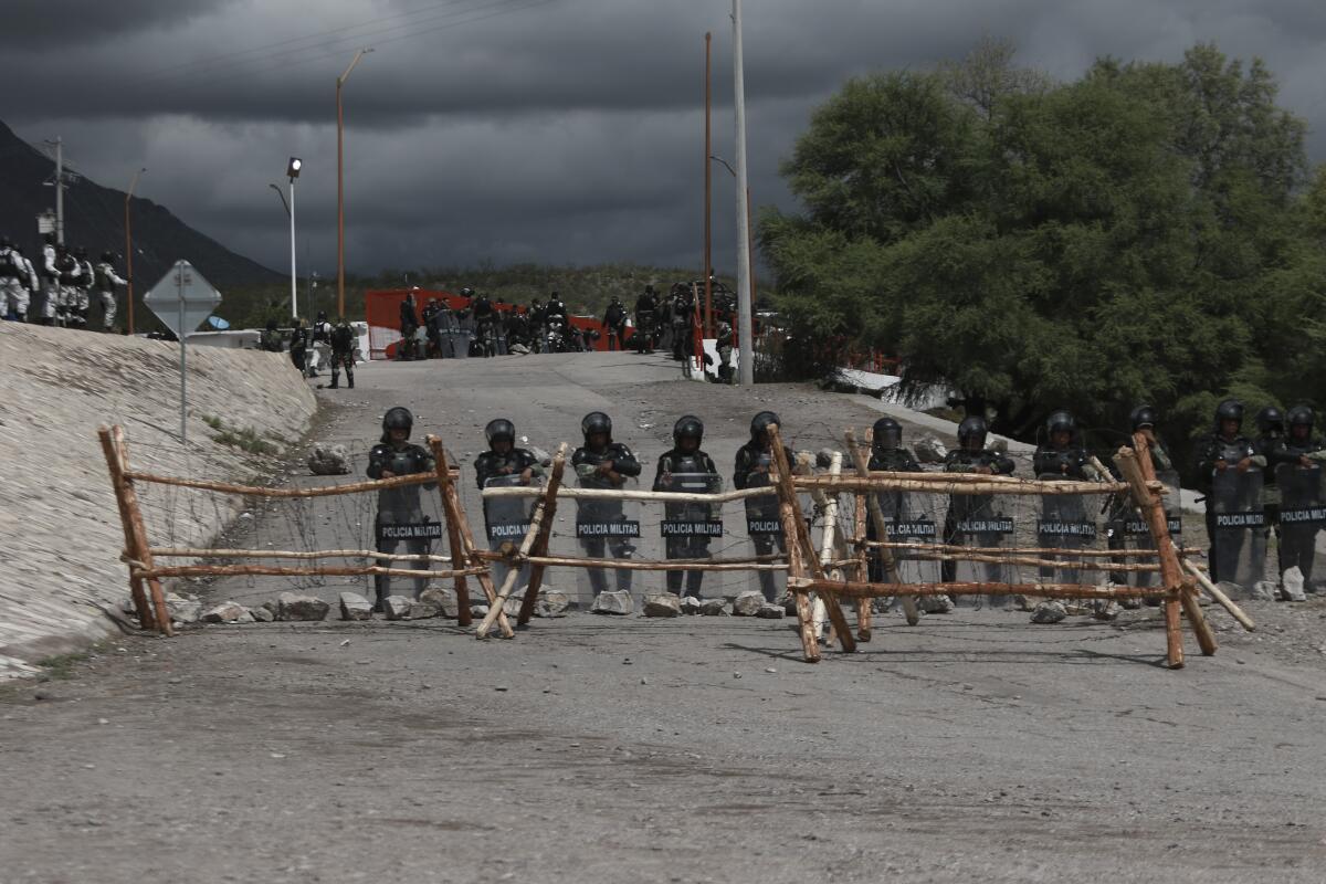 Military police in helmets and riot shields stand behind a wood and barbed-wire barricade on a gravel road