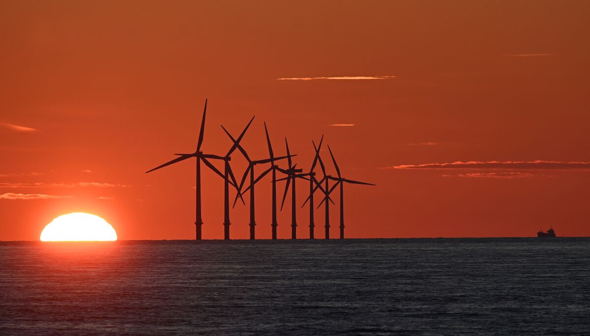 The sun sets behind wind turbines at sea, lending an orange glow to the sky