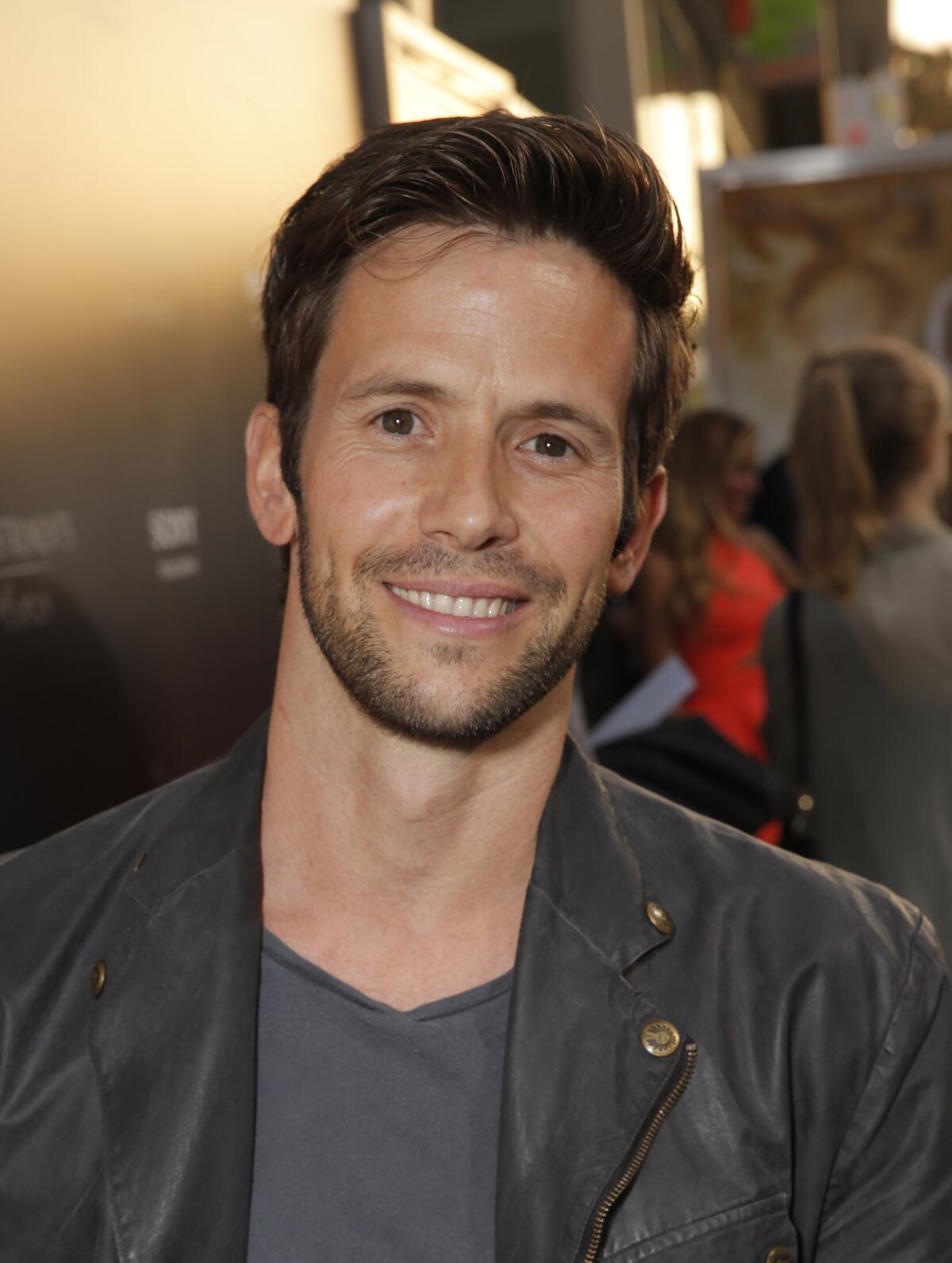 A man with short brown hair and stubble smiling and wearing a black leather jacket and a grey shirt. 