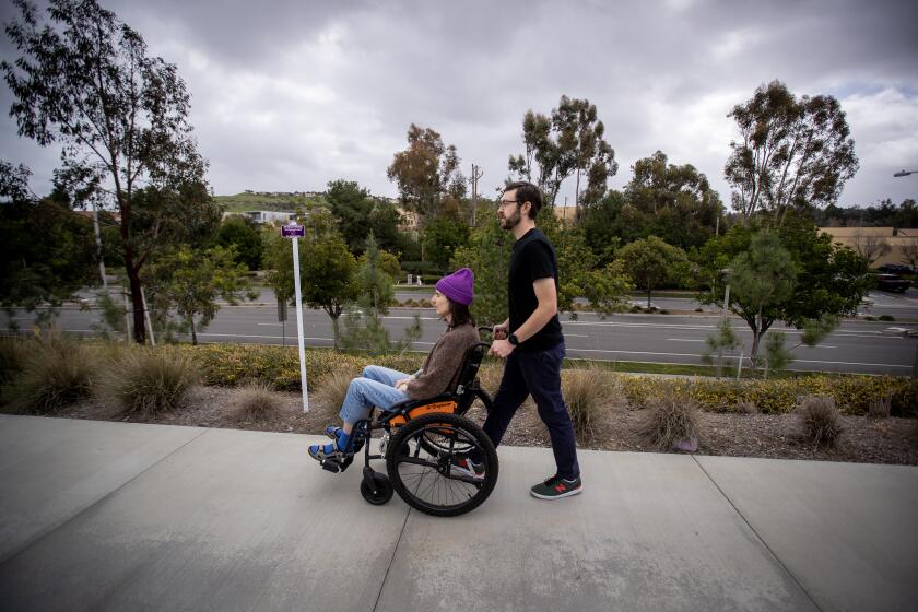 Irvine, CA - February 14: Connor Mayer pushes his wife, Courtney Gavin in a wheelchair as they go outside for fresh air. Courtney got sick with Covid in March 2020 and is now sick with long covid, gets help from her husband doing basic tasks in the home. She is a graphic artist and used to play in a couple bands but is on hold until she recovers at her home with her husband. Photo taken Tuesday, Feb. 14, 2023 in Irvine, CA. (Allen J. Schaben / Los Angeles Times) Courtney is experiencing long covid and the costs (emotionally, physically and financially) this disease has had on their lives. She currently is not working and has severe shortness of breath, needs help doing basic tasks in the home (pushed in a wheelchair, uses a chairlift) and is fatigued easily. Connor works as a professor at UC Irvine and is now her full-time caregiver. He noted he is struggling with balancing the responsibilities of being a caregiver over his career responsibilities (which has resulted in a loss of income).The couple has shared that they have spent over $60k in medical expenses (chair lift, wheelchair, supplements), Courtney plays music and is largely bedridden, but they seem to have a routine where Connor makes her food and sets up everything before he leaves for work to make it as easy as possible for her to eat and take care of herself. Courtney also applied for disability and was denied recently.