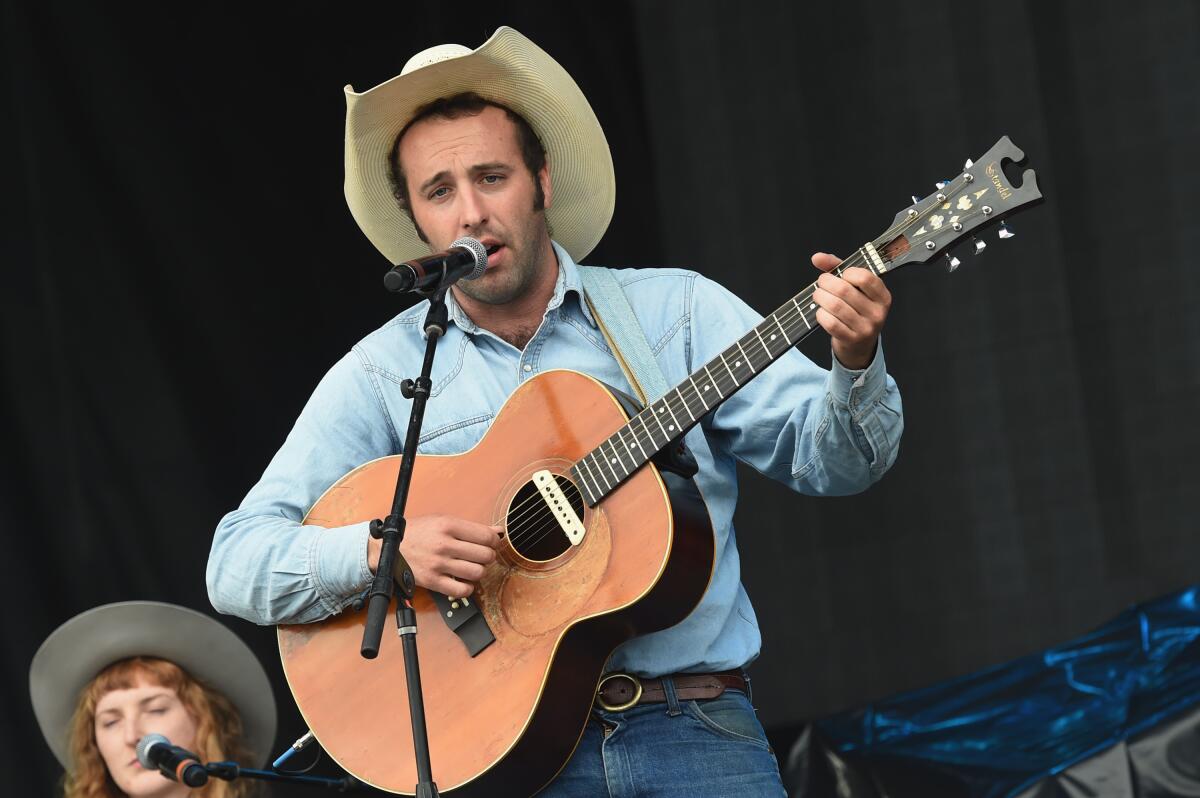 A man with a wide-brimmed hat, holding a guitar and singing into a microphone