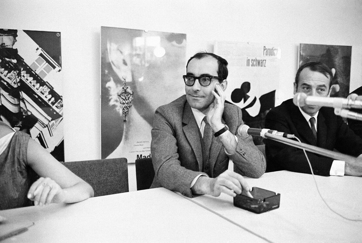 A man in suit and tie and horn-rim glasses sits at a long table, smoking a cigarette, with microphones in the foreground.