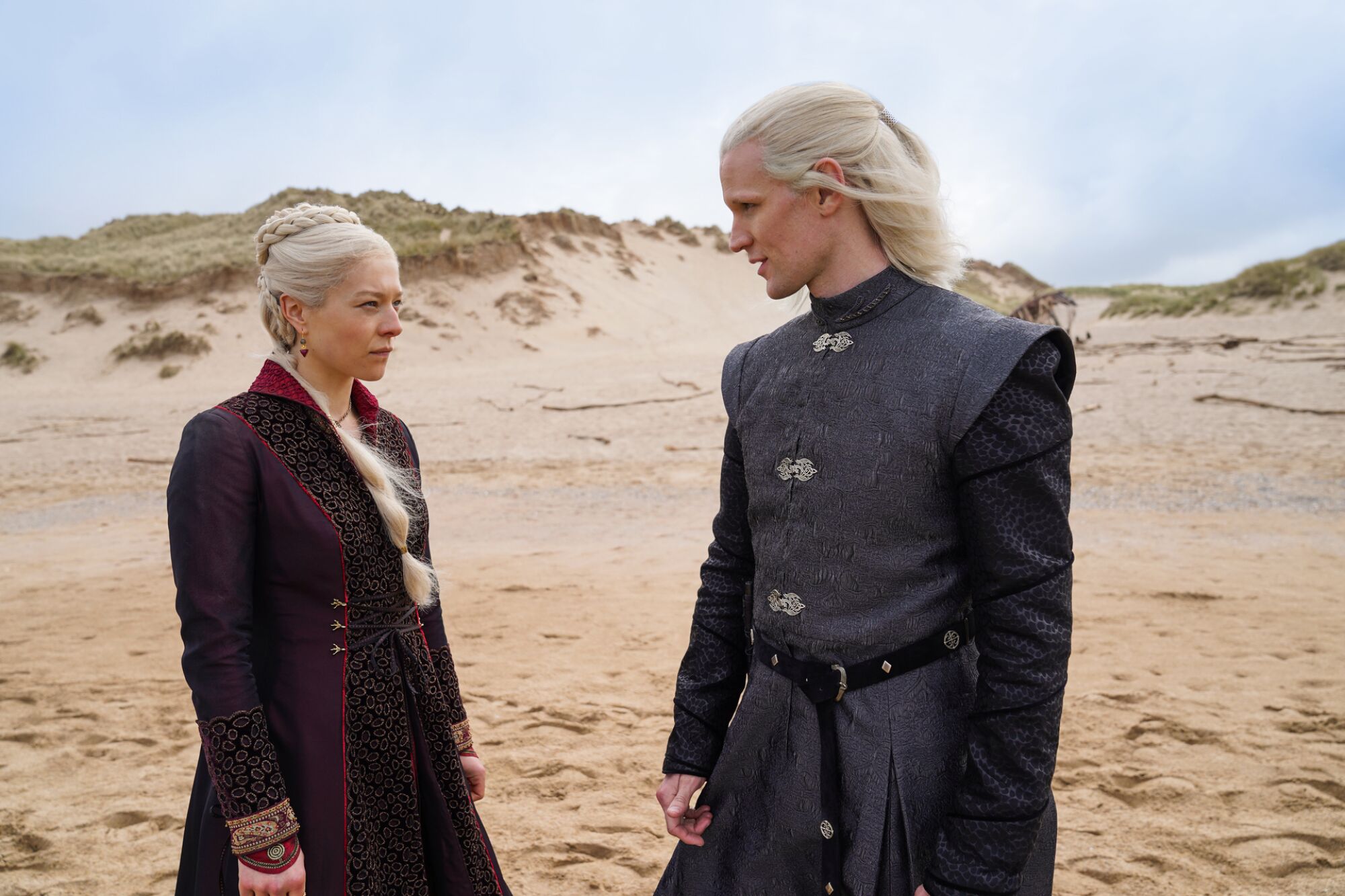 A man and a woman with white-blond hair, wearing medieval costumes, stand on a beach looking at each other.