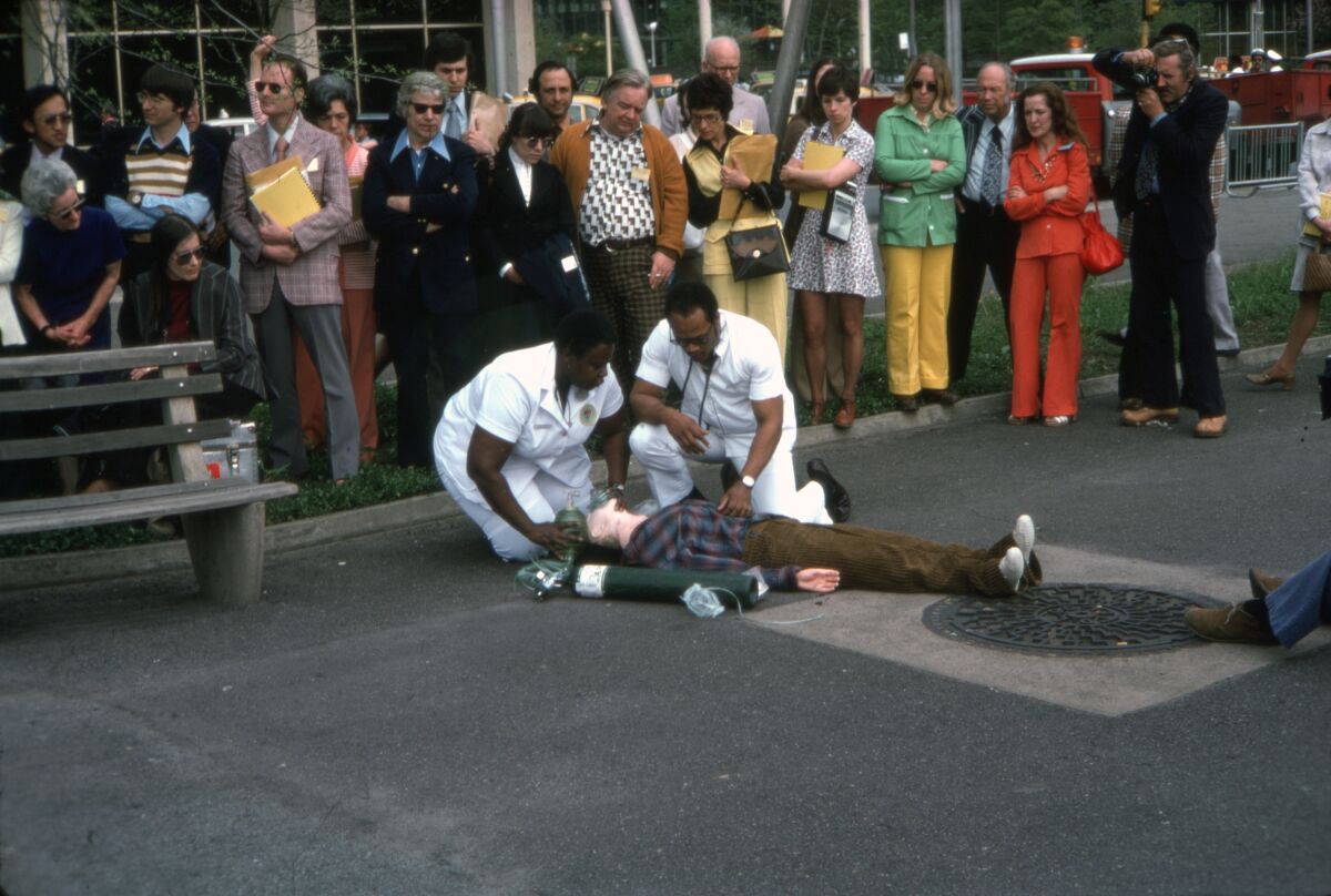 Two uniformed men on their knees tend to a mannequin flat on its back as a crowd observes