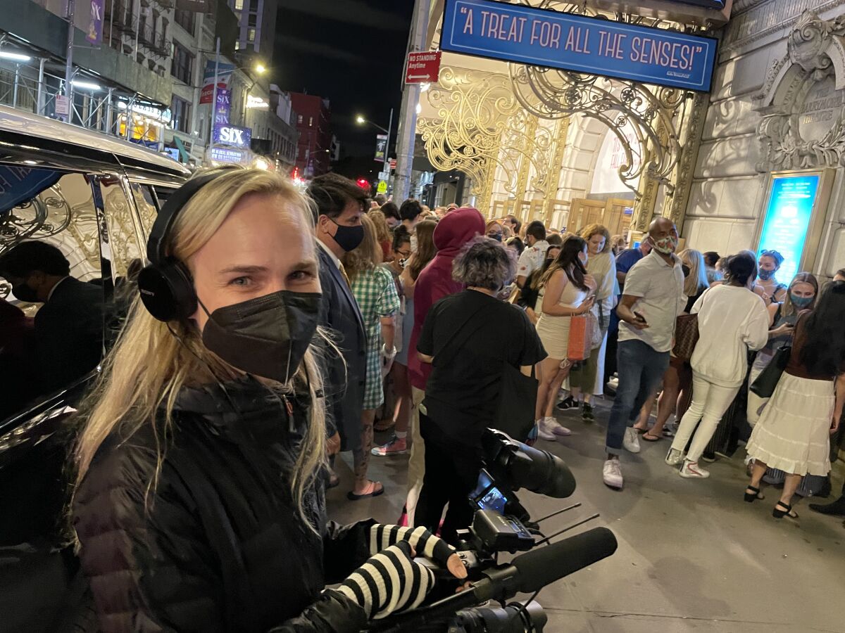 A woman wearing headphones and a face mask holds a microphone outside a Broadway theater.