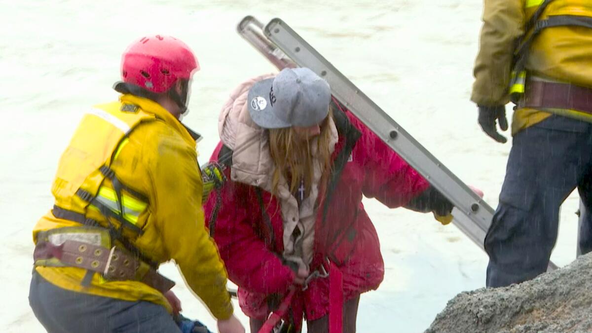 A woman in a pink puffy coat and backward baseball cap is helped near a river by two people in yellow reflective gear.