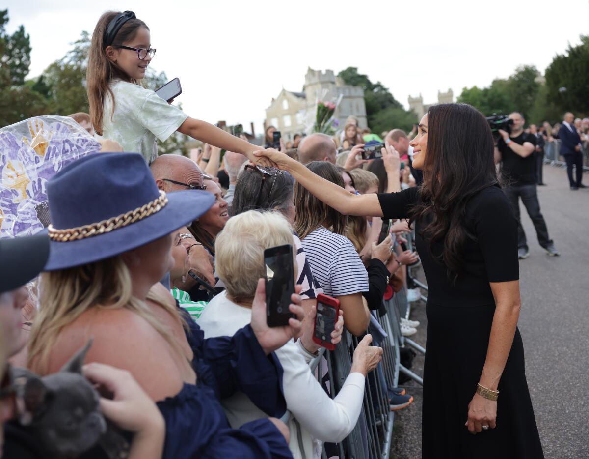 A woman in a black dress reaches over a barrier into a crowd to shake the hands of a girl sitting on a man's shoulders.