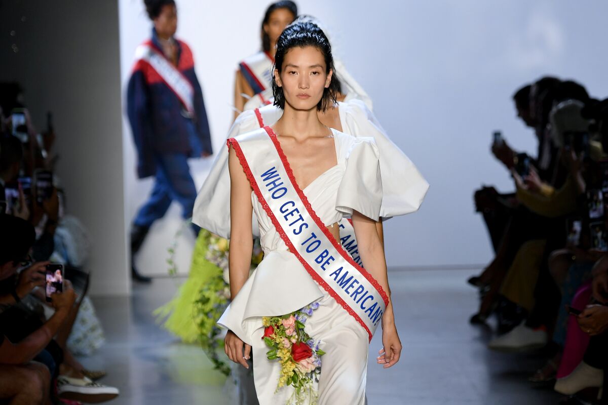 A runway model in a white dress adorned with flowers and a sash that says "Who gets to be American?"