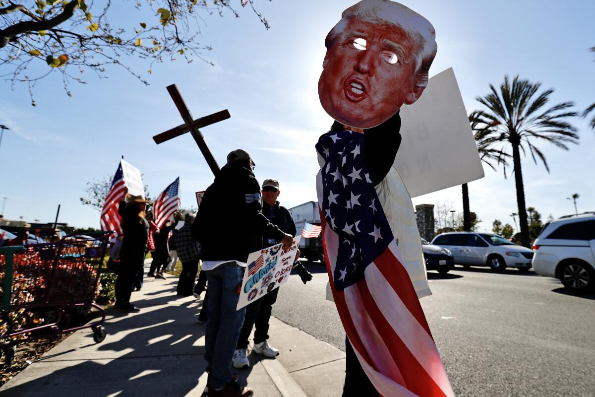 Protesters line a sidewalk. One carries a cross. Another carries a U.S. flag and a large mask of Donald Trump's face.