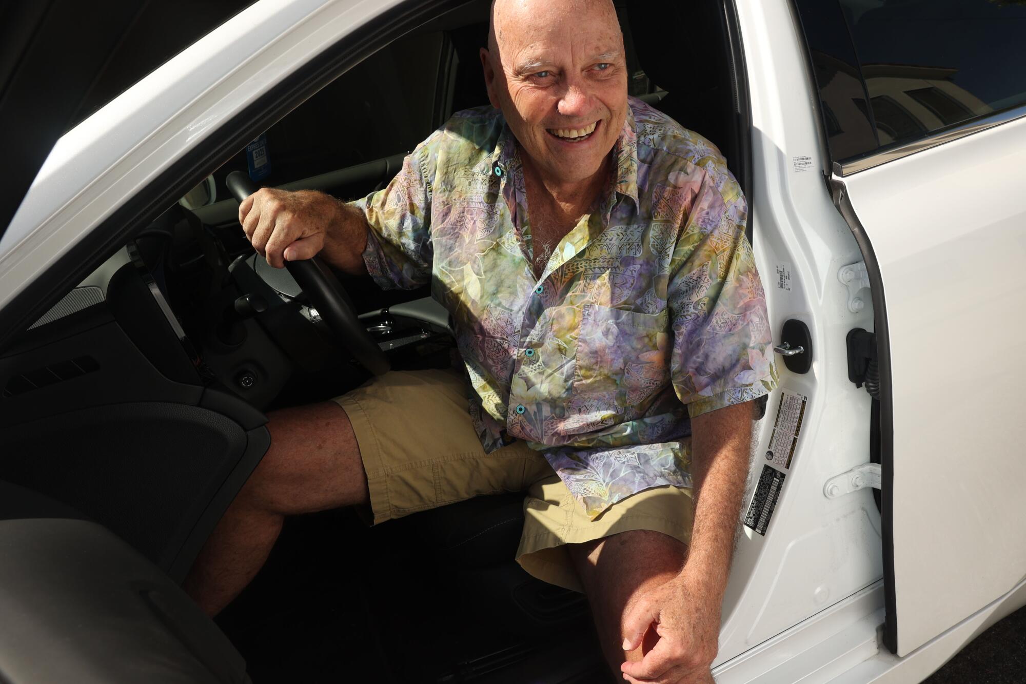 A man with bald head and flower patterned shirt sits in the driver's seat of a car.