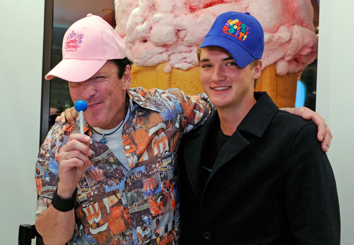 A father and son wearing colorful baseball caps, one holding a lollipop