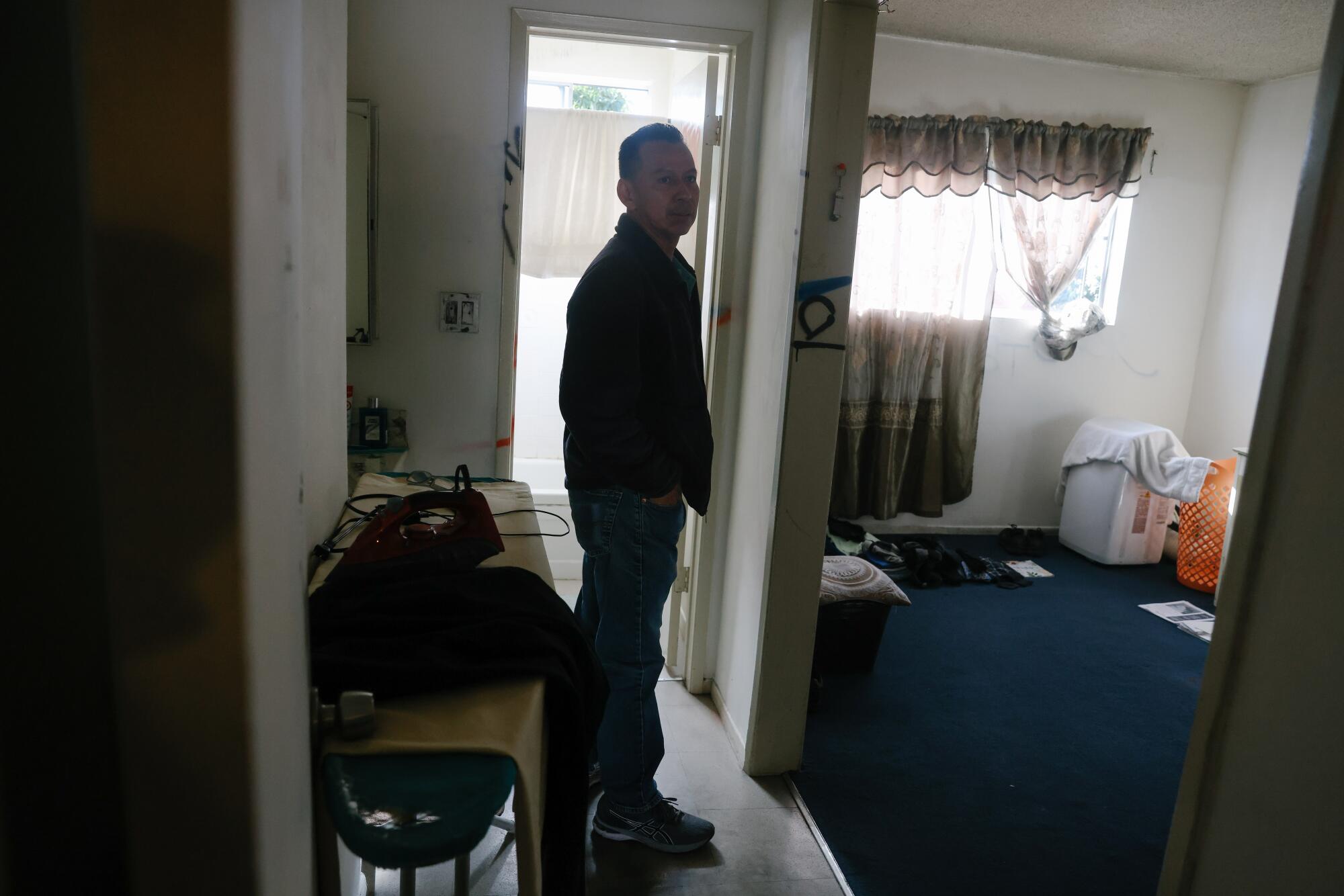 A man standing in an apartment outside a room with a dark-blue carpet 