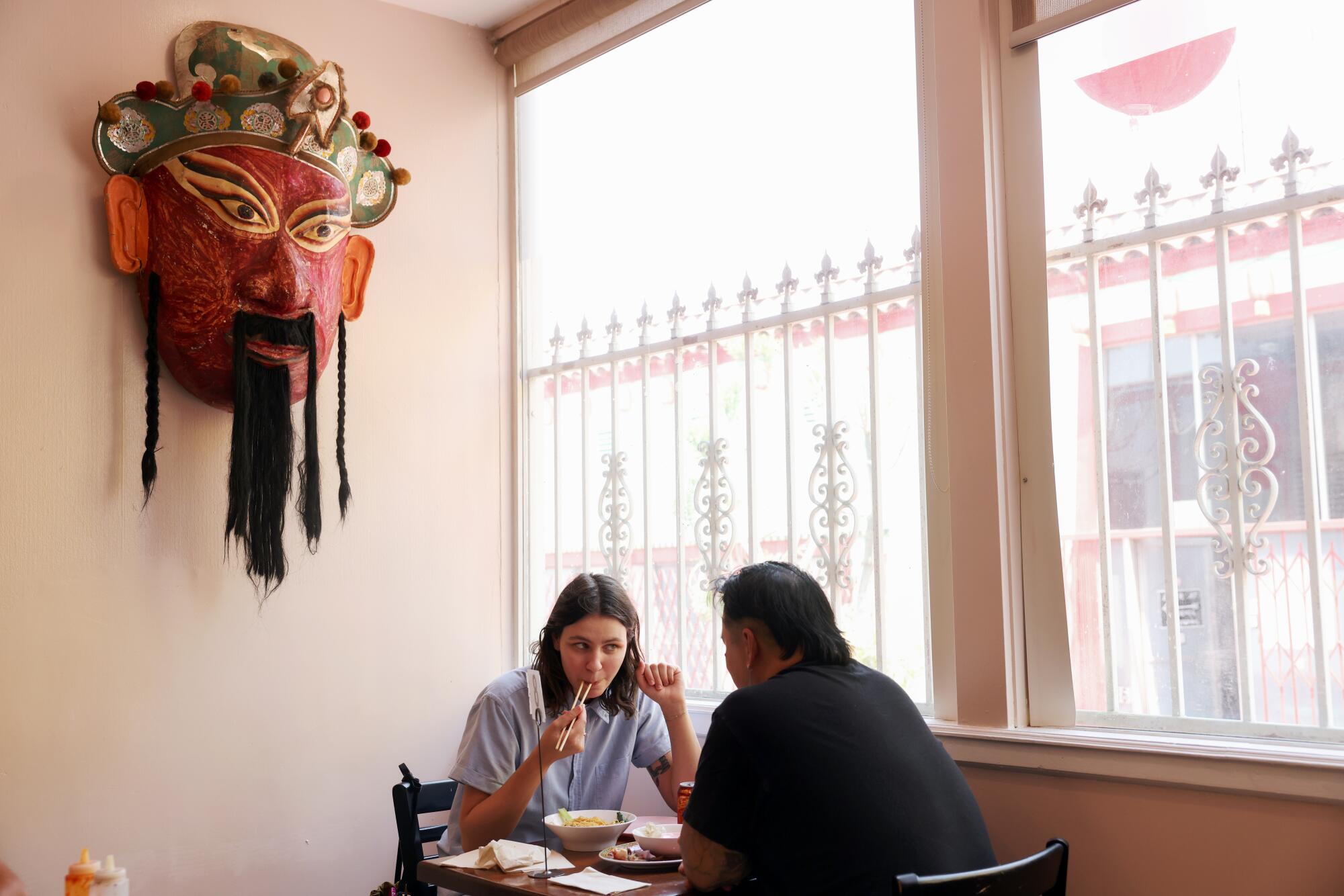 Two people dine in a restaurant next to a window, with a large mask hanging on the wall above them.