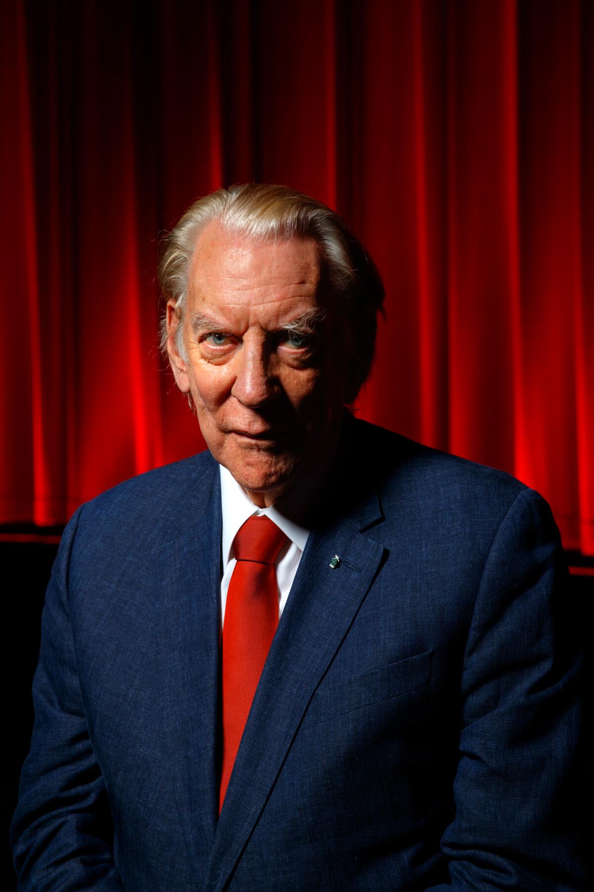 Donald Sutherland, in a blue suit and red tie, stands in front of a red stage curtain.