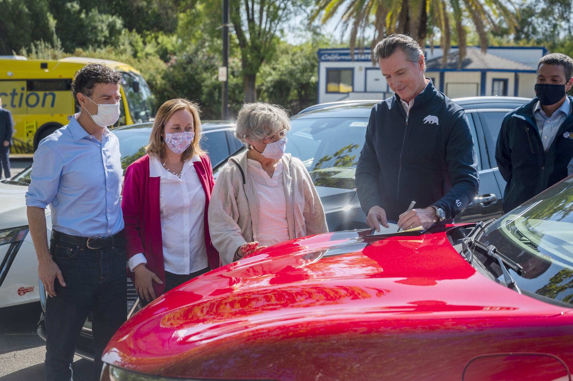 California Gov. Gavin Newsom signs a document on the hood of a shiny red car before onlookers.