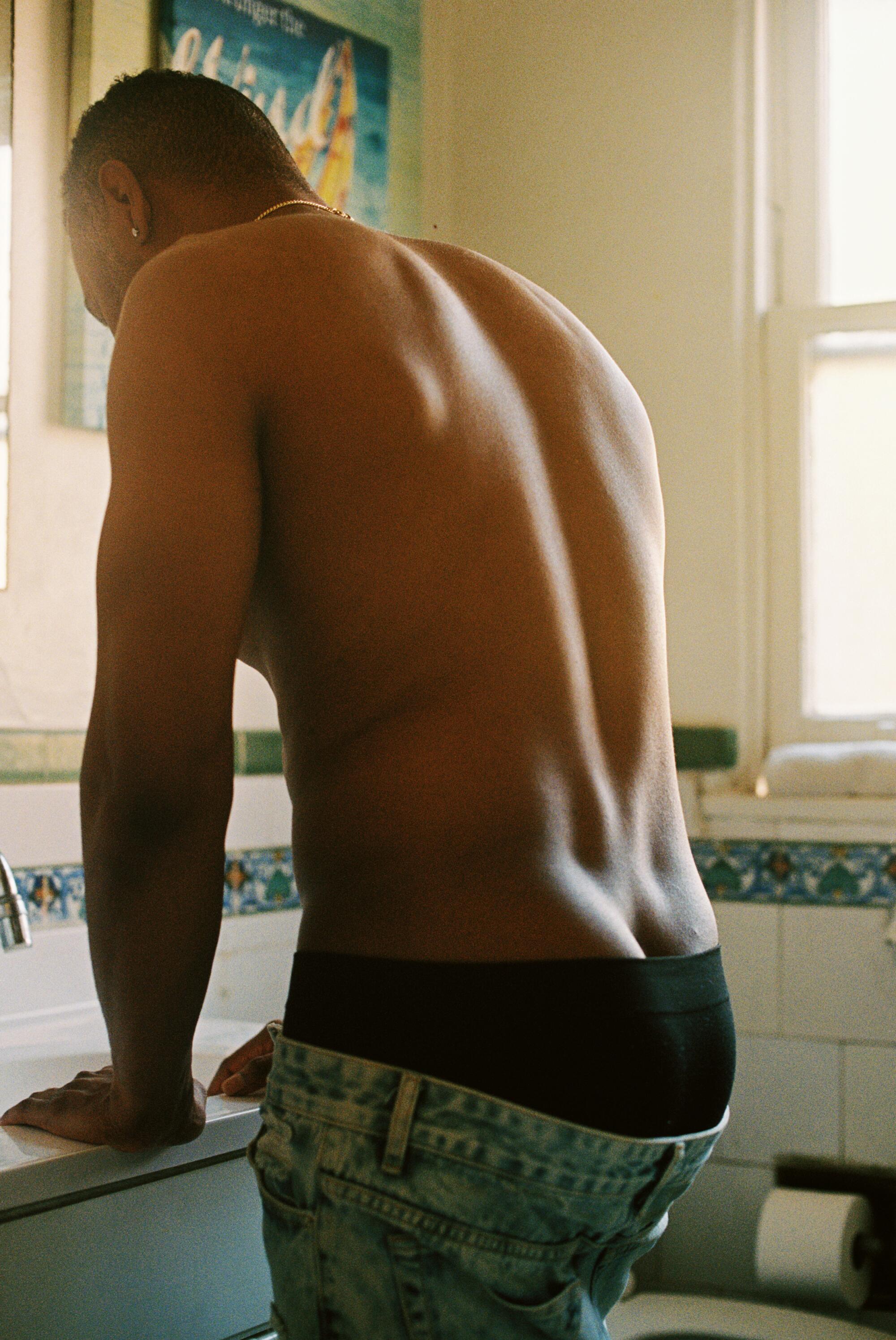 A shirtless man leans over a sink in a bathroom, his underwear showing above his jeans.